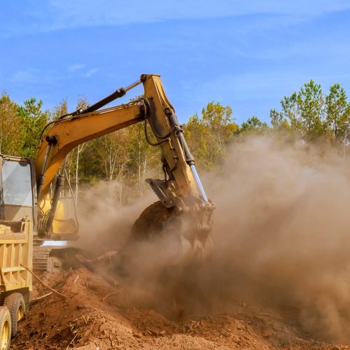 Excavator digging at construction site with dust cloud under blue sky in sunny weather